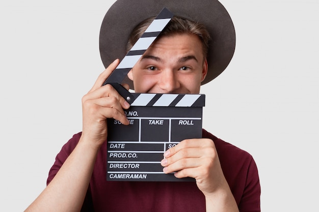 Positive young cameraman holds clapboard near face, has joyful expression, wears hat, prepares for making cutaway, involved in filming, poses on white studio wall. Cinematography concept