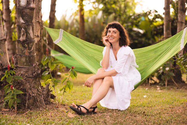 Позитивная молодая кавказская брюнетка носит платье, отдыхает, сидя в гамаке на открытом воздухе летом Концепция уединения с природой