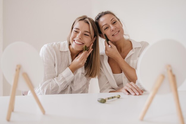 Positive young caucasian women smiling look in mirror while sitting at table using facial scraper. Medical beauty treatments