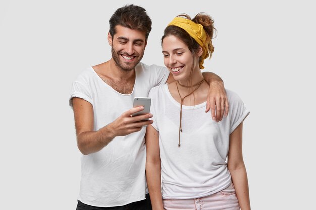 Positive young couple embrace each other, look joyfully at mobile phone, glad to view common photos