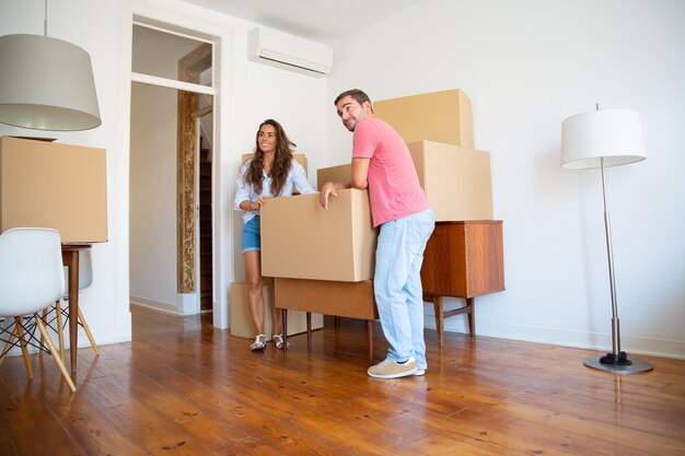 Позитивная молодая пара смотрит на свою новую квартиру, стоя и опираясь на картонные коробки и мебель в помещении