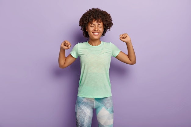 Positive young curly girl stretches arms, feels delighted, does exercises, wears casual t shirt and leggings