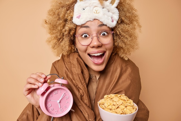 Positive young curly haired woman holds bowl of cereals and alarm clock awakes you for breakfast wears sleepmask wrapped in blanket has good mood isolated over brown background Time to wake up