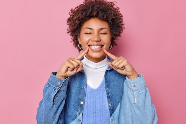 Foto gratuita la giovane donna riccia positiva tiene gli occhi chiusi punti con le dita al sorriso a trentadue denti mostra denti perfetti indossa camicia di jeans esprime emozioni felici isolate su sfondo rosa. concetto di felicità