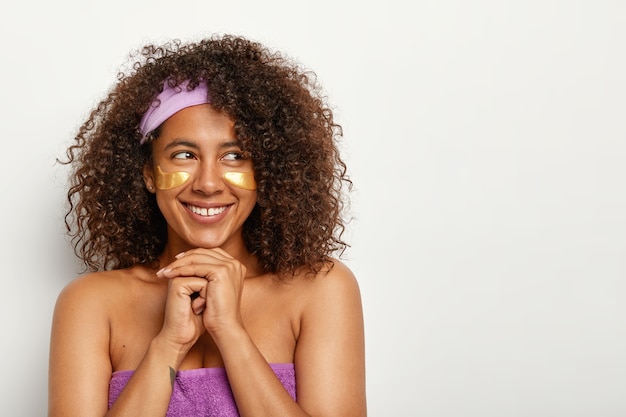 Positive young curly woman looks happily on right, stands with applied under eye patches, keeps hands pressed together under chin, cares about beauty, wears headband, stands bare shoulders indoor