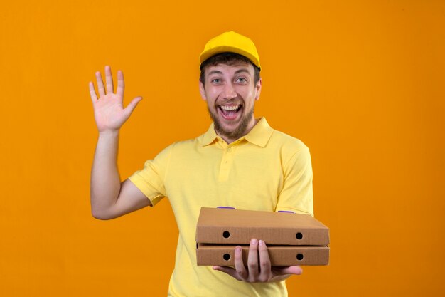 Позитивный молодой курьер в желтой рубашке поло и кепке держит коробки для пиццы, выглядит радостным и делает приветственный жест, машет рукой на изолированном апельсине