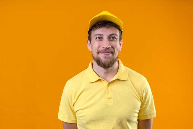 노란색 폴로 셔츠와 모자에 긍정적 인 젊은 배달 남자 격리 된 오렌지에 유쾌 하 게 서 웃 고