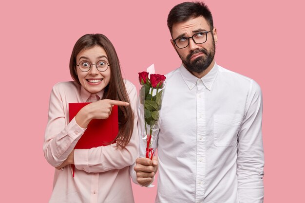 Positive young European woman carries red textbook, points at awkward bearded man in white shirt who feels shy, holds nice bouquet, have love story