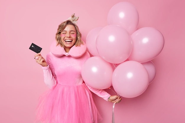 긍정적인 젊은 유럽 여성은 플라스틱 신용 카드로 행복하게 포즈를 취하고 분홍색 배경에서 격리된 미용 절차를 받는 파티 시간을 즐깁니다.