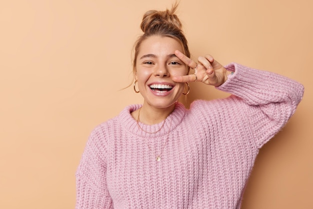 Free photo positive young european woman makes peace sign over eye laughs joyfully enjoys life wears loose knitted sweater isolated over brown studio background. victoryy gesture. body language concept