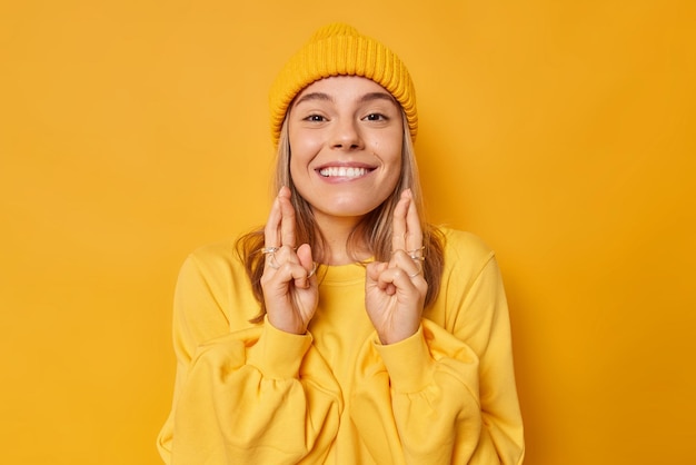 긍정적인 젊은 유럽 여성은 이빨을 가지고 즐겁게 웃고 있으며 행운을 빌며 행운이 모자와 점퍼를 입고 노란색 배경 위에 격리되어 있다고 믿습니다. 신체 언어 개념