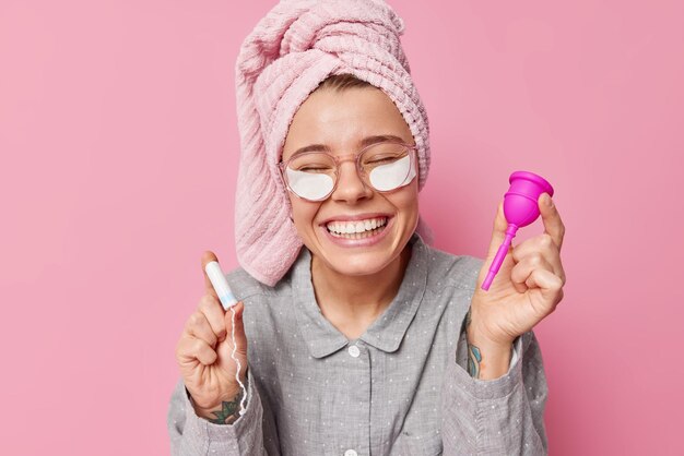 Positive young European woman wears comfortable pajama and wrapped towel on head holds tampon and menstruation cup during menses isolated over pink background applies beauty patches under eyes