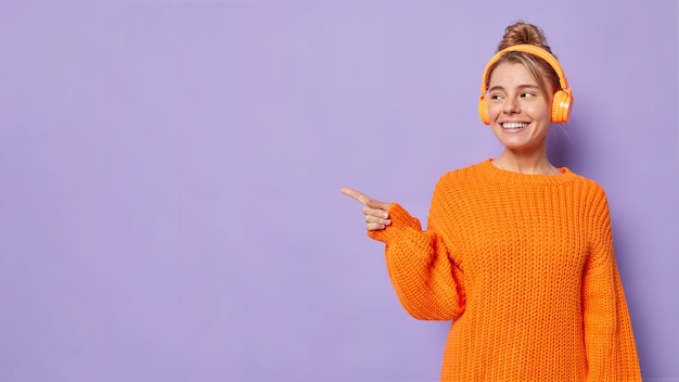 Positive young European woman with combed hair listens audio track via headphones points away at blank copy space wears knitted sweater shows place for your advertisement attracts your attention