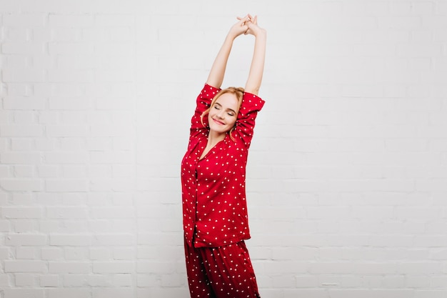 Free photo positive young female model stretching on white wall. beautiful fair-haired woman in sleepwear relaxing in morning.