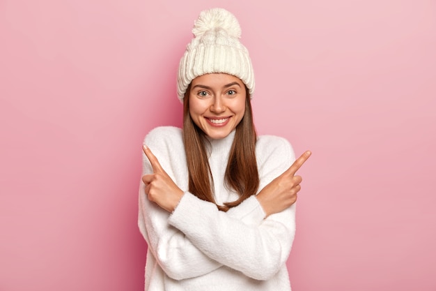 Positive young girl with straight hair, crosses arms and points in both sides, wears fashionable white hat and jumper, chooses between two items