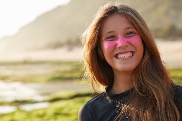 Positive young glad European woman with toothy smile, has protective zinc mask on face which blocks sun rays, wears diving suit for surfing, poses outdoor against blurred coastline wall.