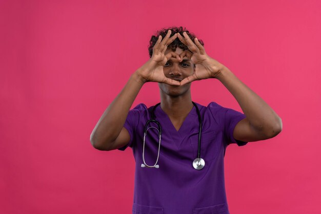 Un giovane medico dalla carnagione scura bello positivo con capelli ricci che indossa l'uniforme viola con lo stetoscopio che si tiene per mano sulla forma del cuore