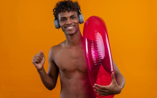 A positive young handsome dark-skinned man with curly hair in headphones enjoying music while holding inflatable pool ring 