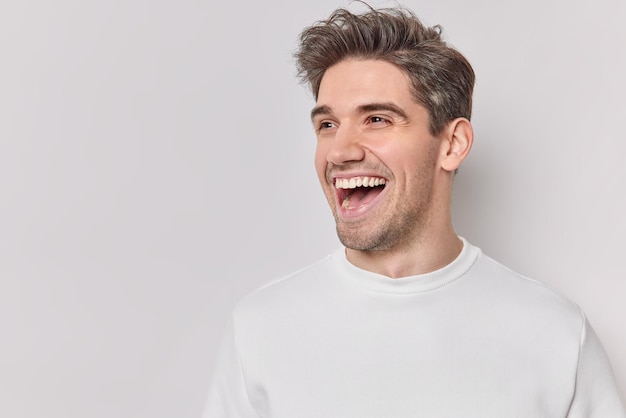 Positive young handsome man giggles positively laughs at something funny looks with excited joyful expression dressed casually isolated over white background blank copy space for your advert