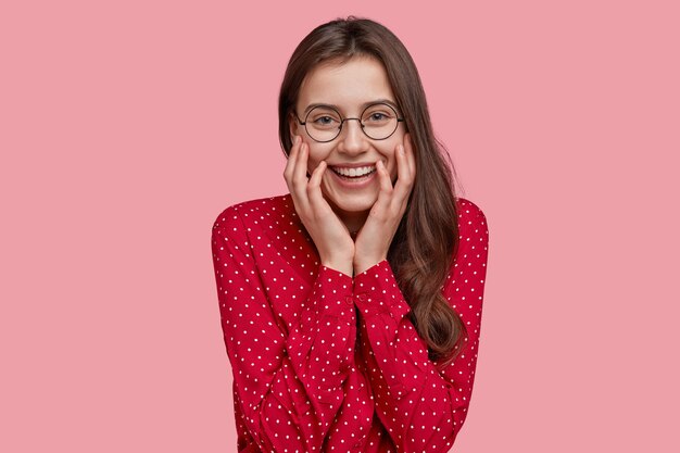 Positive young lady with gentle smile, wears spectacles, fashionable shirt, demonstrates natural beauty