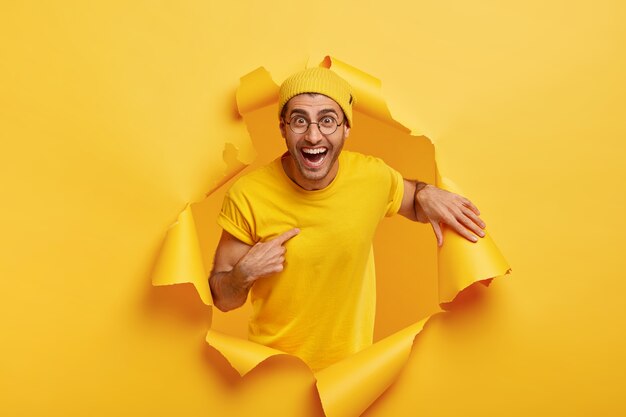 Positive young male model stands in hole of ripped paper background, points at himself, asks about choosing his own canditature