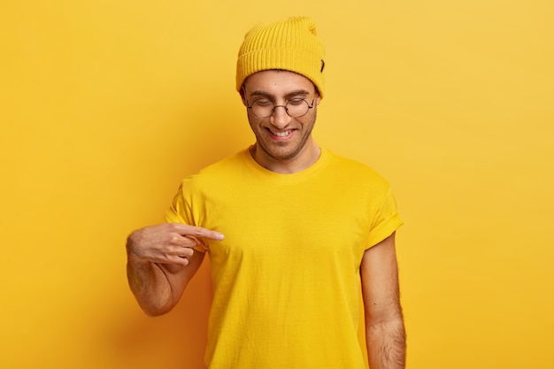 Il giovane maschio positivo indica lo spazio vuoto della maglietta, mostra lo spazio per il tuo design o logo, sorride volentieri, indossa gli occhiali, vestito giallo, concentrato verso il basso