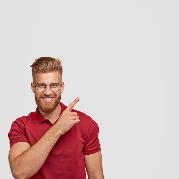 Positive young man being satisfied with results of work, has friendly pleasant smile, points upwards with fore finger, wears casual clothes, stands in against white wall