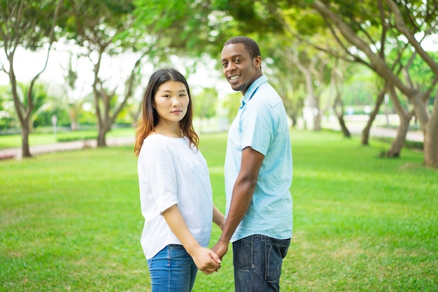 公園で歩いている間、手を持っているポジティブな若い多民族のカップル