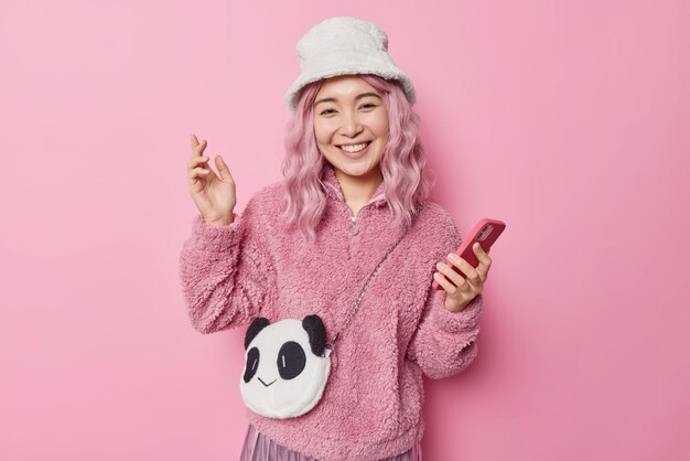 Positive young pretty Asian woman with pink hair wears hat and jacket carries panda bag holds smartphone smiles positively glad to receive message from boyfriend isolated over pink background