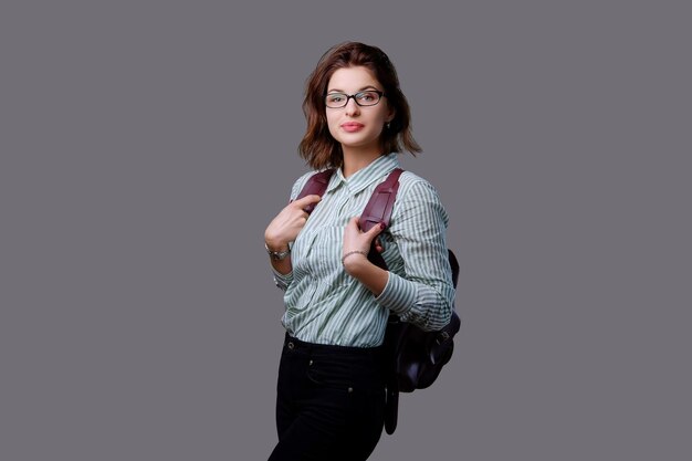 Positive young traveler female with leather backpack isolated on grey background.