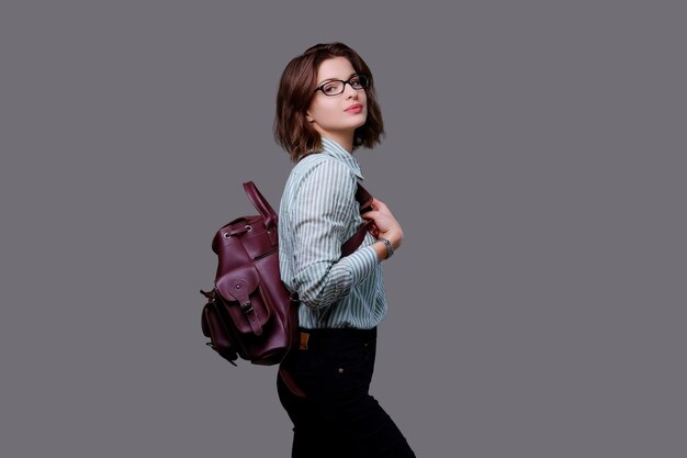 Positive young traveler female with leather backpack isolated on grey background.