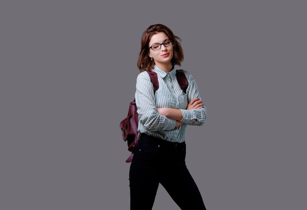 Positive young traveler female with leather backpack isolated on grey background.