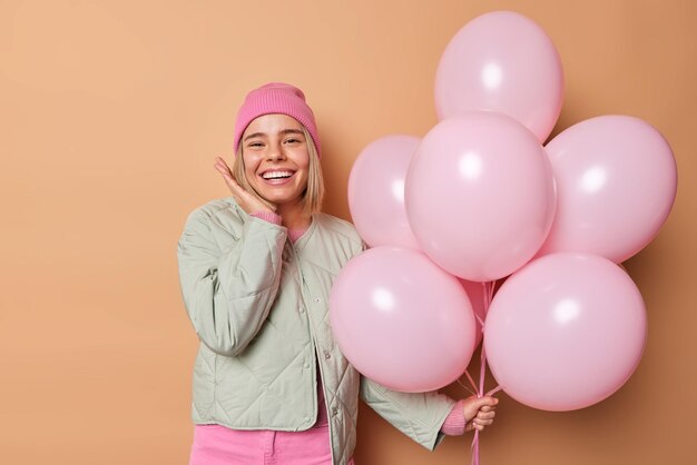 Positive young woman celebrates special occasion wears hat and jacket smiles happily holds bunch of inflated balloons comes to congratulate friend with birthday isolated over brown background