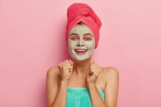 Positive young woman clenches fists with joy, applies clay mask on face, wears pink towel on head, wants to have natural beauty, models indoor, smiles broadly, expresses happiness. Wellbeing