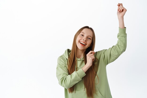 Positive young woman dancing and having fun, wearing casual hoodie, celebrating with dance, smiling cheerful, white wall