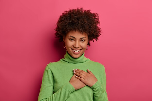 positive young woman feels thankful, keeps hands pressed to heart, wears green turtleneck, smiles positively, looks with tender smile, isolated over pink bright wall. thank you very much