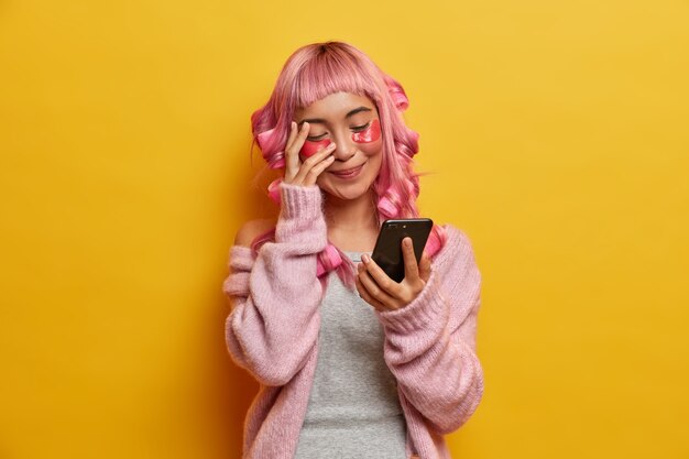 Positive young woman giggles positively and looks at smartphone display, reads funny news, has long pink hair, makes hairstyle, cares about skin