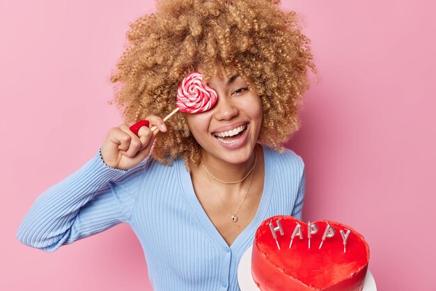 Positive young woman has sugar addiction and sweet tooth holds heart shaped cake lollipop over eye smiles broadly wears blue jumper has good mood isolated over pink background Confectionery