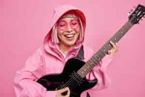 Free photo positive young woman keen on music plays favorite music holds electric guitar smiles happily being in good mood wears anorak with hood on head trendy sunglasses