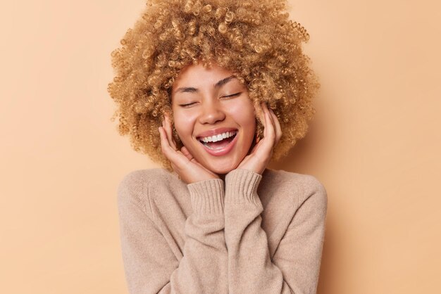 Positive young woman keeps eyes closed touches face gently smiles broadly has perfect white teeth dressed in casual jumper isolated over beige background. Happy emotions and feelings concept