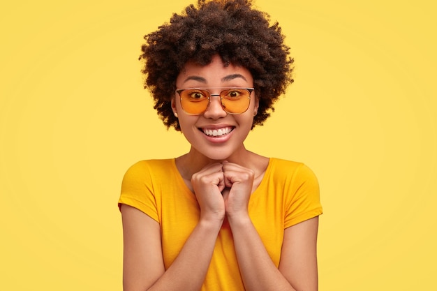 Foto gratuita la giovane donna positiva tiene le mani unite sotto il mento, ha un ampio sorriso, denti bianchi perfetti, vestita con una maglietta luminosa