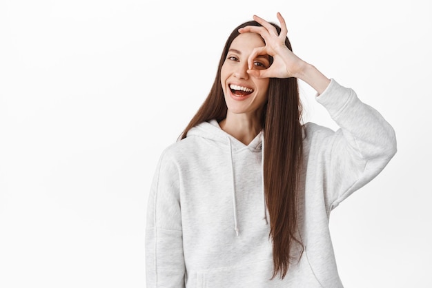 Positive young woman say yes shows okay sign on eye and smiling make OK agree gesture praise perfect excellent choice satisfied by something good standing over white background
