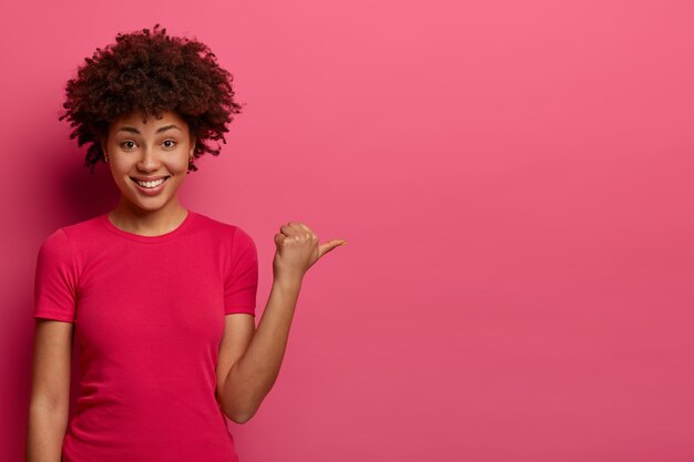 Positive young woman shop assistant helps customer to find dressing room, advertises product on sale, points thumb aside, wears casual t shirt, smiles gladfully, isolated on pink wall.