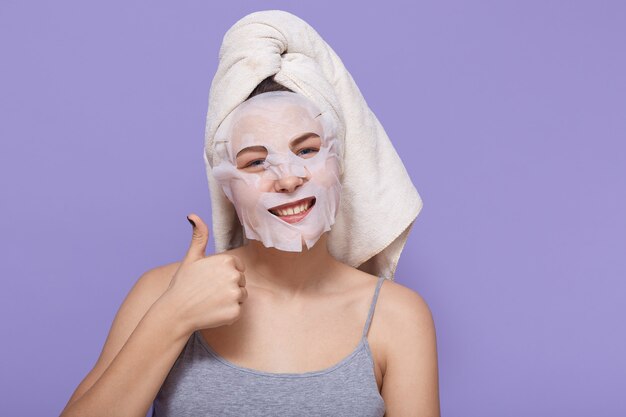 Positive young woman shows her big thumb up while posing with cosmetic mask on her face