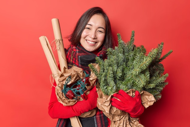 緑のトウヒの枝と花輪が赤い背景の上に隔離された新年とクリスマスの前に家を飾るつもりで冬の服のポーズで幸せに身を包んだポジティブな若い女性の笑顔。