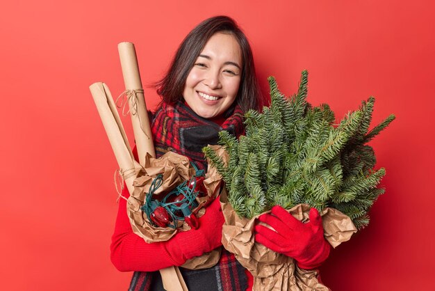 緑のトウヒの枝と花輪が赤い背景の上に隔離された新年とクリスマスの前に家を飾るつもりで冬の服のポーズで幸せに身を包んだポジティブな若い女性の笑顔。