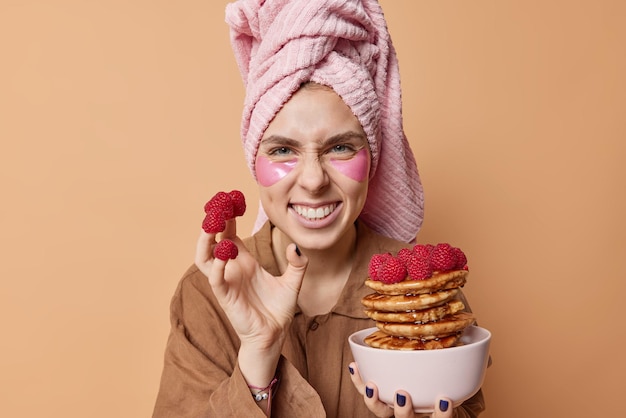 Positive young woman smirks face and shows white teeth has fun while eating delicious breakfast has raspberries on fingers wears wrapped towel on head pajama applies beauty patches under eyes