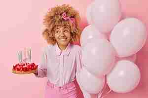 Бесплатное фото Позитивная молодая женщина в модной блузке и джинсах позирует с вкусным клубничным тортом и кучей воздушных шаров празднует особый случай, изолированный на розовом фоне празднование дня рождения