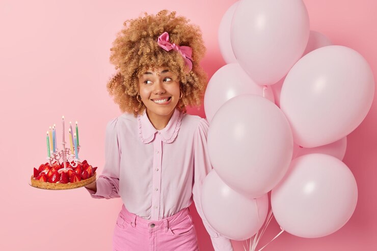 Free Photo positive young woman wears fashionable blouse and jeans poses with delicious strawberry cake and bunch of balloons celebrates special occasion isolated over pink background birthday celebration