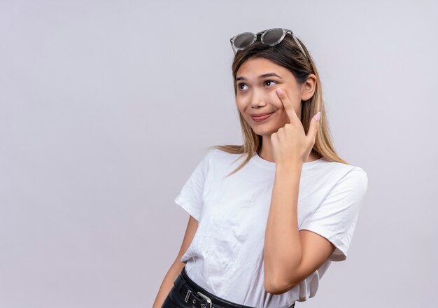 A positive young woman in white t-shirt in sunglasses smiling and pointing with index finger at cheek on a white wall